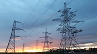 Metal transmission towers forming an overhead line. Contractors in PPE stand atop a gantry suspended from a tower's arm.