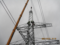 Contractors in PPE stand atop a metal transmission tower, the uppermost section of the tower is suspended from a crane arm.  A date in the corner reads "2021/10/31 10:10".