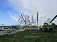 Sections of metal transmission tower laying on the ground, contractors in PPE stand amongst the sections and near construction vehicles.