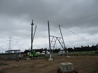 One side of the base of a metal transmission tower. Contractors in PPE stand around a crane nearby.