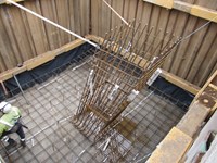 An excavated area with metal supports, inside the area a diagonal hollow cuboid of rebar extends upwards.