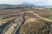 An aerial view of construction vehicles on a dirt road within a rural region. Mountains are present in the distance.