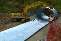 Contractors in PPE lifting a metal sheet into place, adding to a tubular corrugated metal structure on a construction site.