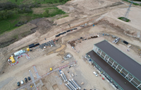 An aerial view of a construction site. A metal framed building with a black roof is prominent.