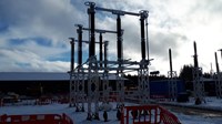 Substation infrastructure surrounded by construction barriers, construction vehicles are parked in the background. Thick snow sits on the ground.