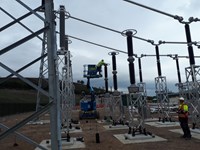 Substation infrastructure in a substation under construction, a contractor in PPE and a harness stands nearby. A mobile elevator raises another contractor up to some of the infrastructure.