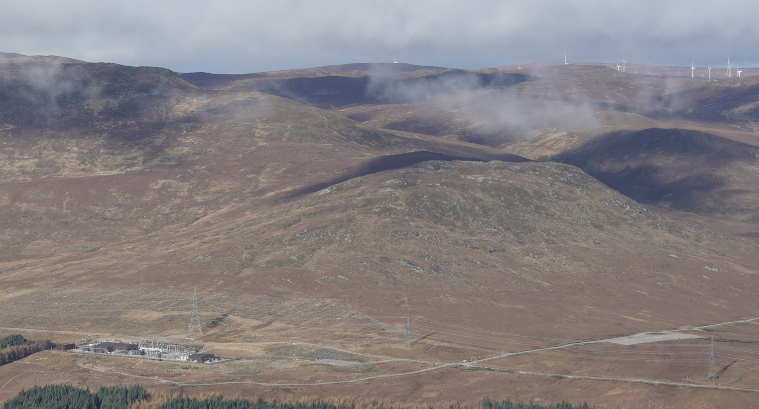 A substation in a valley with wind turbines in the hills.