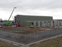 Construction vehicles including a crane adding sections of green material to the outside of a metal framed building surrounded by substation infrastructure.