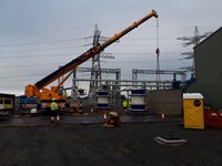 An orange crane attached by chains to a container on the ground, contracters in various locations around this as well as substation infrastructure.