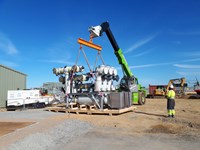 A section of transmission infrastructure resting on pallets, attached by chains to a crane beam.