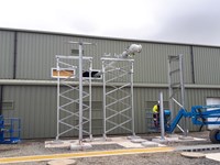 An engineered pipe emerging from a green-walled building, supported by metal frames with concrete bases. A contracter nearby stands on a mobile lift vehicle.