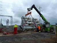 A section of transmission infrastructure is suspended above the ground by chains using a crane vehicle. Two contractors in PPE stand nearby in contact with the section.