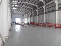 The interior of a metal framed building, a group of contractors in PPE stand at the far end near a large open entrance.