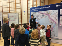Primary school aged children with an SSEN Transmission employee stand in a hall next to a large poster titled "Orkney Infrastructure Project" "The South Section." Other smaller posters are present along a wall.