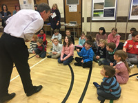 An SSEN Transmission employee speaks to primary school aged children seated on a hall floor. Staff and information posters are present along a wall behind them.
