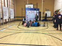 Primary school aged children with an SSEN Transmission employee seated on a hall floor next to a large poster titled "Orkney Infrastructure Project" "The South Section." Other smaller posters are present along a wall.