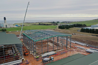 Metal framed buildings under construction on a reddish muddy construction site. Two cranes are present reaching over the buildings. Concrete industrial towers are visible in the background.