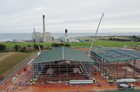 Metal framed buildings under construction on a reddish muddy construction site. Two cranes are present reaching over the buildings. Concrete industrial towers are visible in the background.