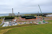 A partially completed metal transmission tower, the uppermost section of which is suspended from a crane. An industrial site under construction is visible in the background.