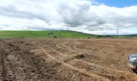 A dirt area, in the background a grassy hill is crossed by transmission towers forming and overhead line.