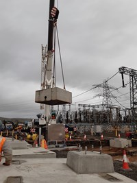 A concrete cuboid suspended from a crane by chains, hanging over concrete slabs and surrounded by substation infrastructure.