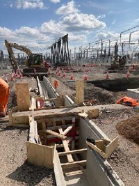 A concrete lined trench with wooden supports. An excavator and substation is visible in the background.