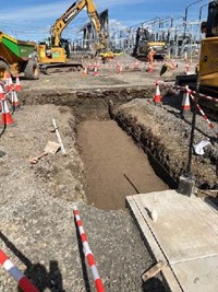 A dirt trench, construction vehicles are present in the background with substation infrastructure behind them.