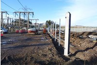 Metal fence posts in a dirt area, substation infrastructure is present on one side of the fenceposts. A contractor in PPE and a construction vehicle are nearby.