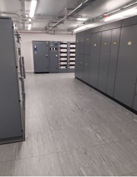 Several large dark-grey metal cuboids along the wall of a florescent-lit room with a grey material floor comprised of squares of material.