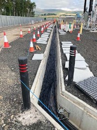 A concrete-lined trench in a substation under construction. Large amounts of cables lie along the base of the trench. A series of concrete slabs are laid along the ground next to the trench.