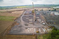 An aerial view of a substation section under construction. A crane and other construction vehicles are present, and transmission infrastructure is visible to the side of the construction area.