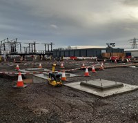 Concrete sections embedded in a construction site floor. In the background transmission infrastructure is visible, as well as construction supplies and metal-walled buildings.