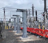 Metal vertical structures embedded in concrete, in the background transmission infrastructure is present.