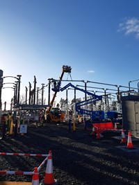 A section of substation infrastructure suspended from a construction vehicle. A contractor in PPE inspects the structure on a mobile elevated platform.