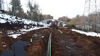 Green and black cylinders laid in a dirt trench, surrounded by light snow with a construction vehicle in the distance.