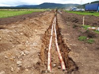 A dirt trench. Tape marked "DANGER ELECTRIC CABLE BELOW" runs in two lines the length of the trench.