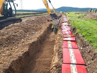 An excavator pours earth onto black and green cylinders at the bottom of a trench. Nearby are red slabs marked "DANGER ELECTRIC CABLE BELOW".