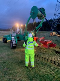 A contractor stands in PPE in front of a construction vehicle with a wire spool and plough attachment.