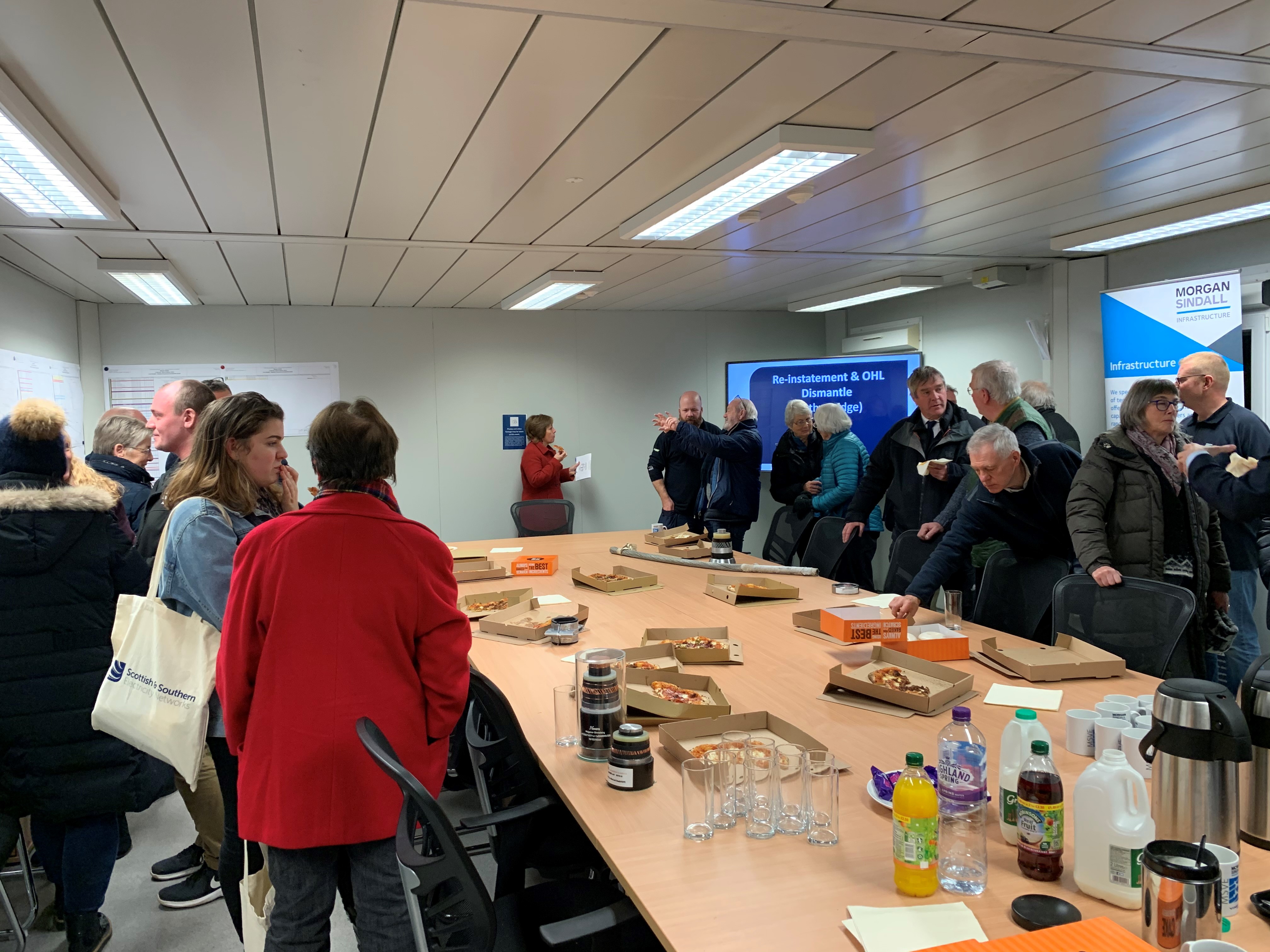 Members of the public stand surround  a table with food and refreshments, posters are present on the walls of the room.