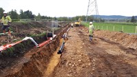 Contractors in PPE next to an excavated trench, an excavator is visible in the background.