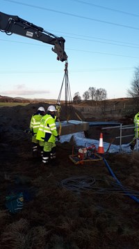 A section of concrete suspended by chains in the air, cables extend from each end of the section. Behind there is an excavated area lined with a material. Contractors surround the suspended section.