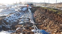 An excavated trench with repeating sets of metal structures along it, several thick black cables lie in the trench and are covered by sandbags at one end.