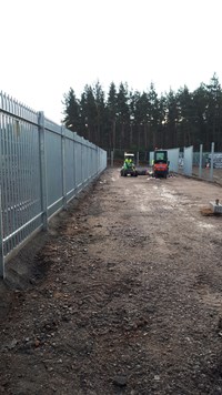 A metal fence surrounding a cleared dirt area. Construction vehicles driven by contractors in PPE are present.