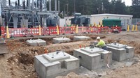 A contractor in PPE stands among several waist-high concrete structures embedded into an excavated area on a construction site. Substation infrastructure is present in the background.