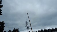 Contractors in PPE sit atop a metal transmission tower under construction. The uppermost section of the tower is suspended from a crane arm.