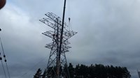 Contractors in PPE sit atop a metal transmission tower under construction. An arm section of the tower is suspended from a crane arm.