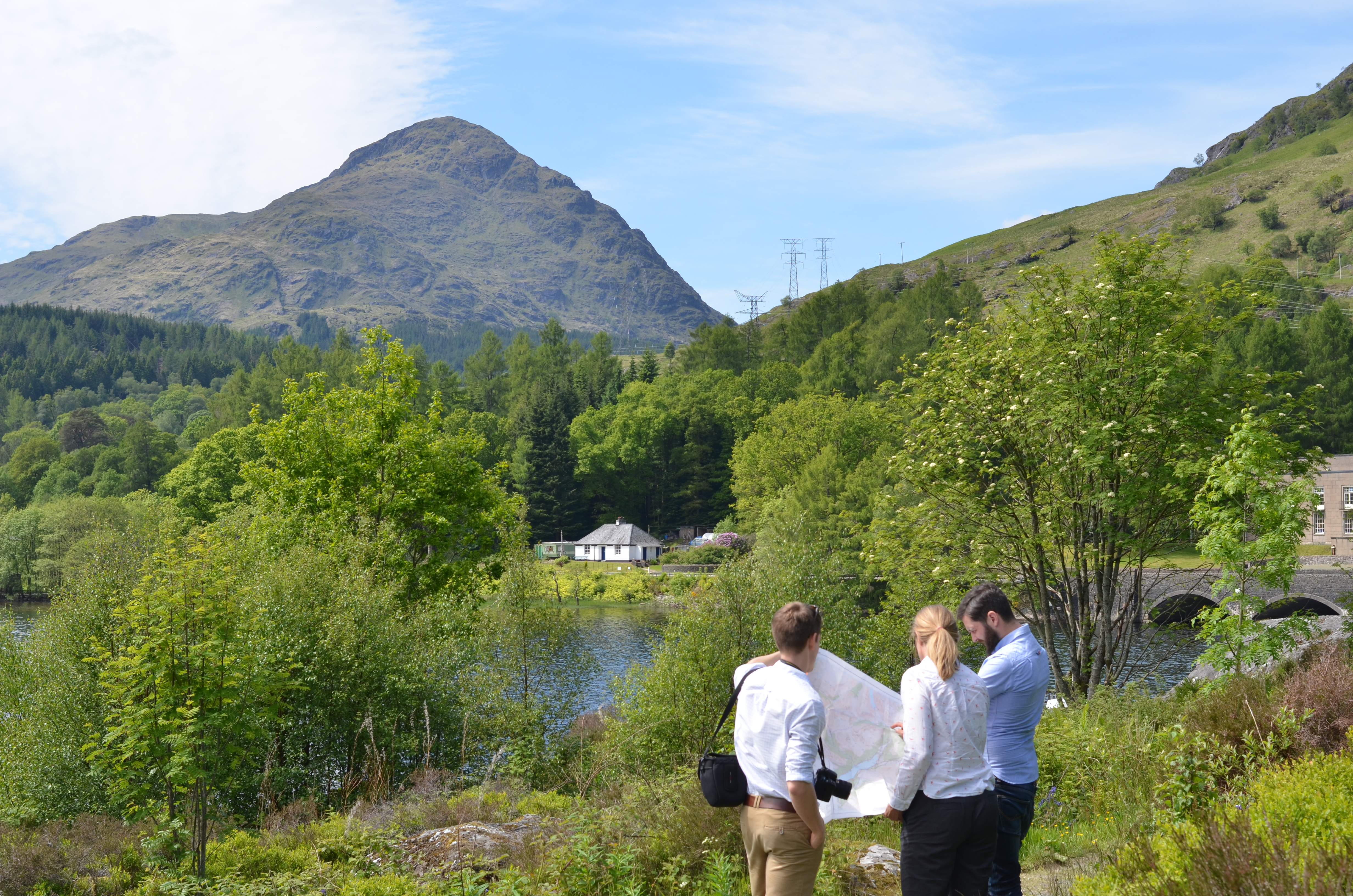 A vegetated area surrounding a body of water. A stone bridge crosses the water and there is a large rocky hill in the distance as well as metal transmission towers.