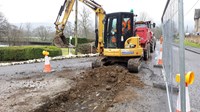 A contractor in a construction vehicle on a surfaced road, in front of the vehicle is a freshly dug trench.