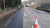 A newly surfaced rectangle of road, in the distance a construction vehicle with an excavation attachment is present.