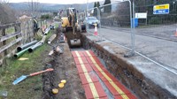 A trench next to a road, a construction vehicle with an excavation attachment is present at the end of the trench. The floor of the trench is lined with  red sections of material and yellow tape.